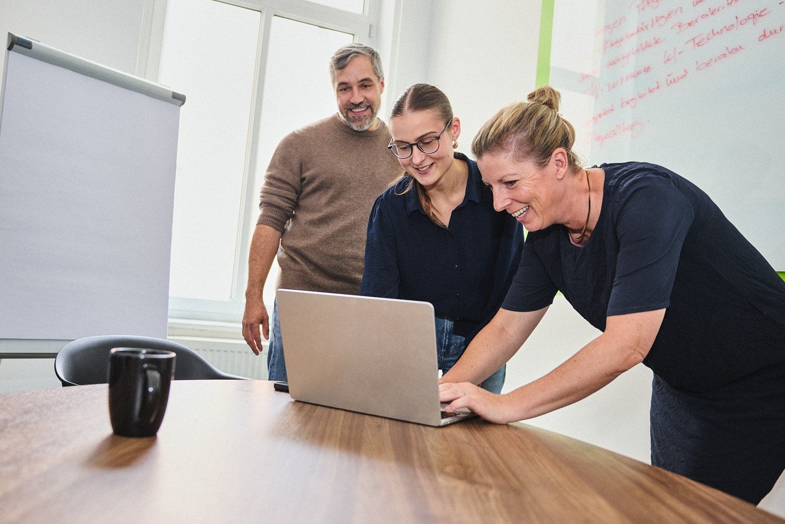 Drei Teammitglieder schauen lachend gemeinsam auf einen Laptop im Besprechungsraum